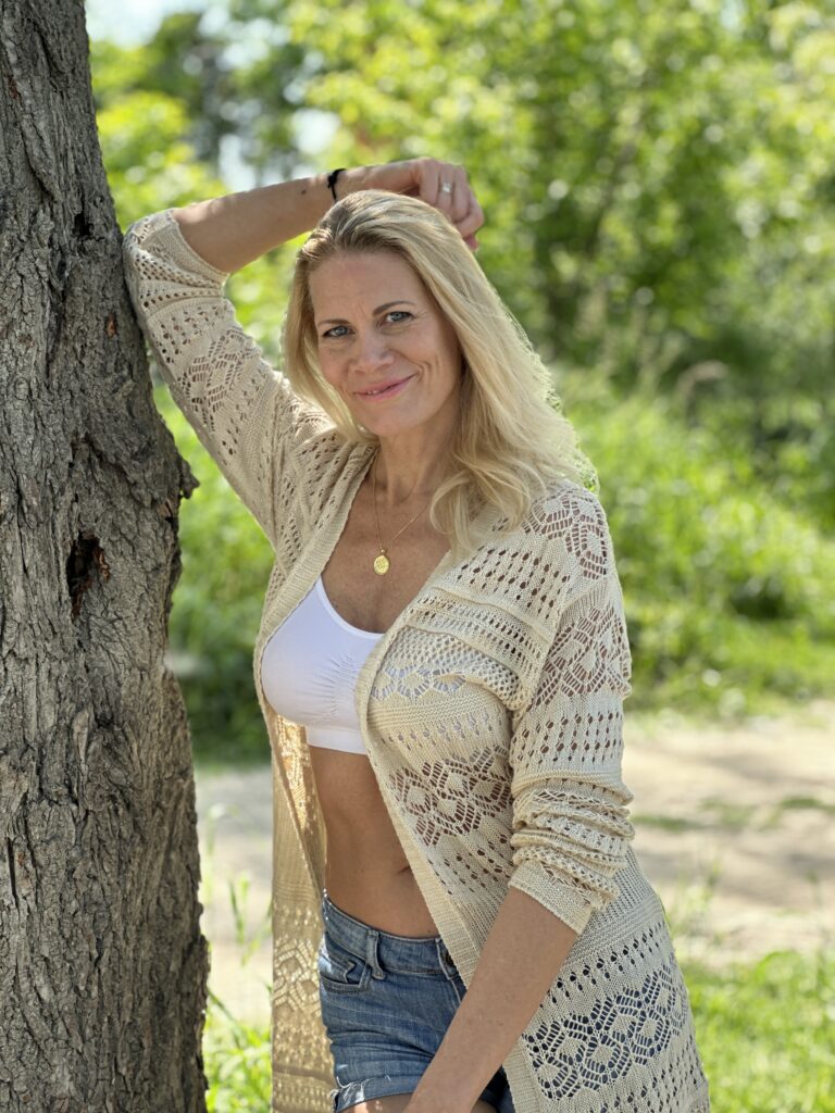 sophies.model leaned against a tree posing for a lifestyle shoot wearing denim shorts, white bra and a knitted cardigan.