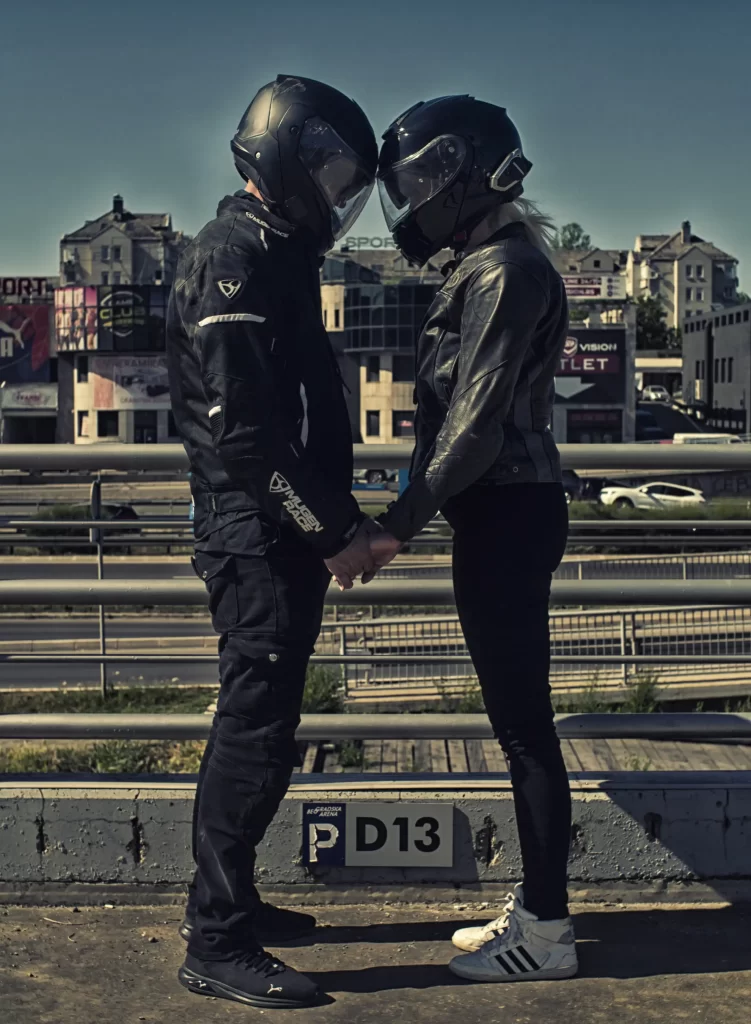 Full shot of SophieS and Rag in full black motorcycle gear facing each other holding hands in a rooftop parking lot.