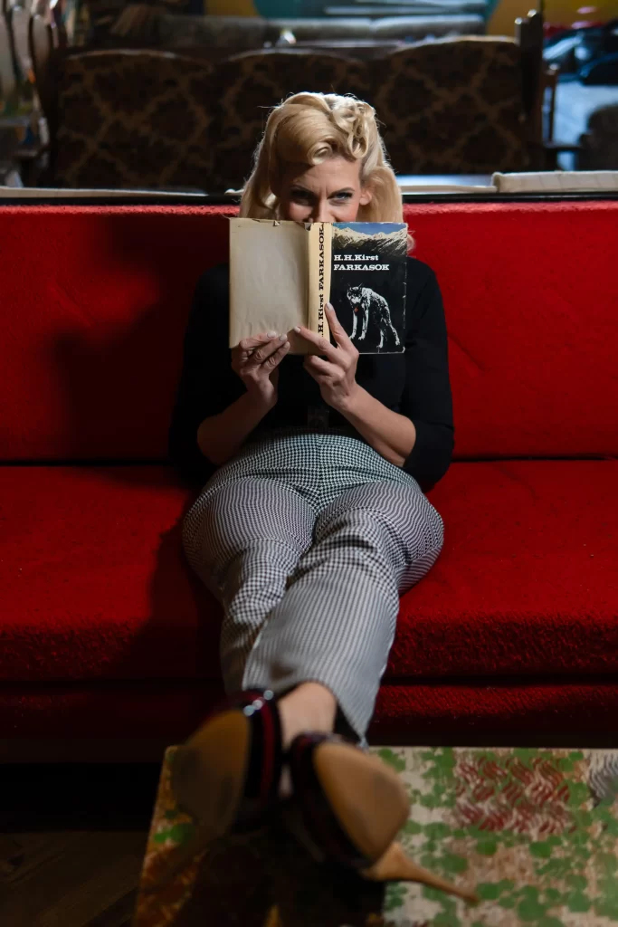 SophieS sitting on a red couch holding a book partially covering her face with a cheeky smile in her eyes.