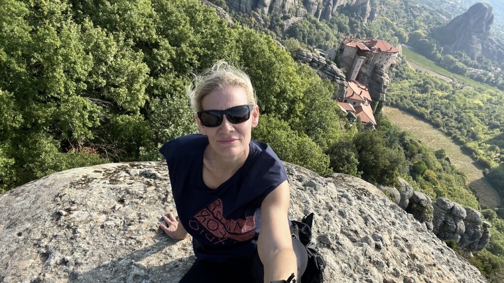 Zsofi sitting on top of the Meteora with a monastery on another peak in the background.