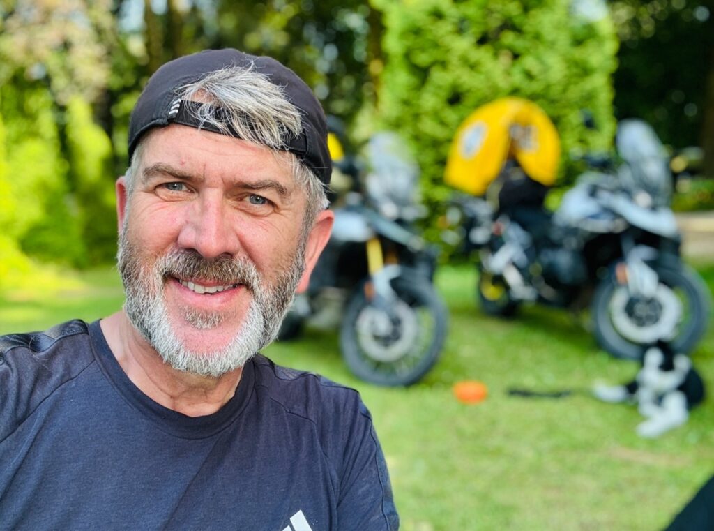 Ken selfije a német kempingben, a motorkkal a háttérben, Molly pedig fekszik a motork mellett.